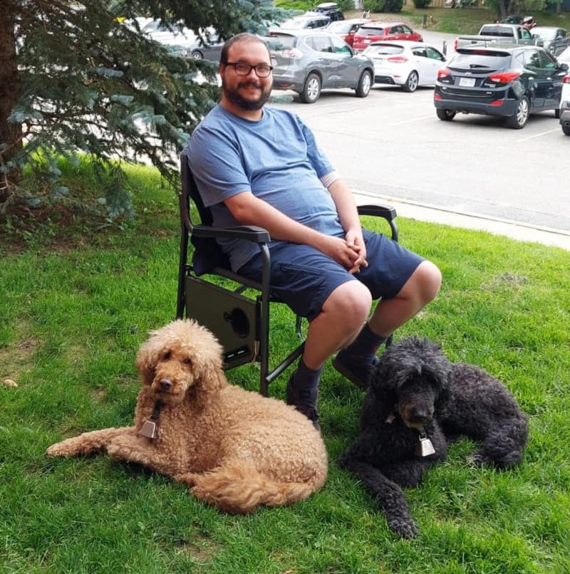 A man is dressed in a t-shirt and shorts, leaning in a camp chair with a golden labradoodle and black standard poodle at his feet